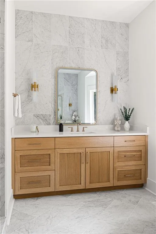 Custom light wood vanity with metallic hardware and a solid-surface countertop, floor-to-ceiling stone wall tile, wall-mounted metallic sconces, and rectangular-tiled stone flooring