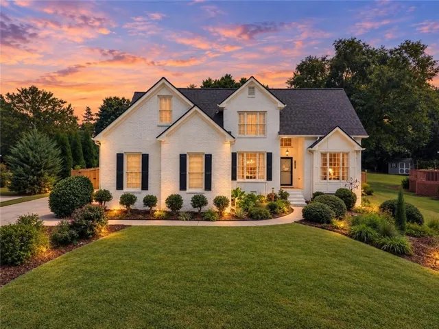 White brick exterior with black shutters, dark shingle roof, bay window, and manicured landscaping with pathway lighting