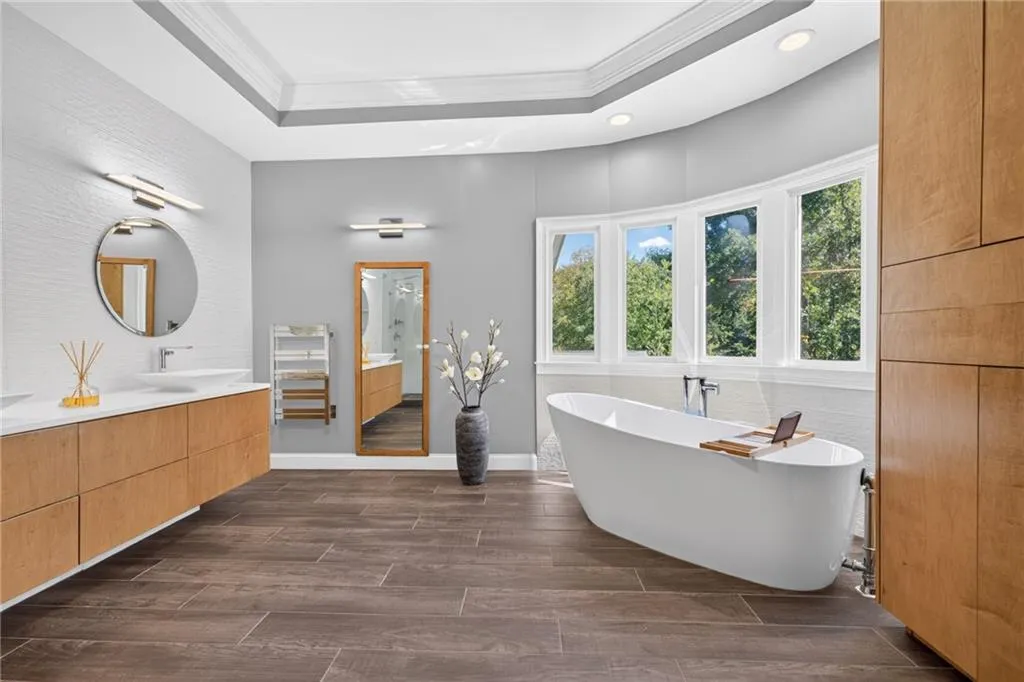 Floating wood-finish vanity with rectangular vessel sinks and stone-surfaced countertop, freestanding soaking tub, wood-finish tile flooring, tray ceiling with recessed lighting, and bay window arrangement