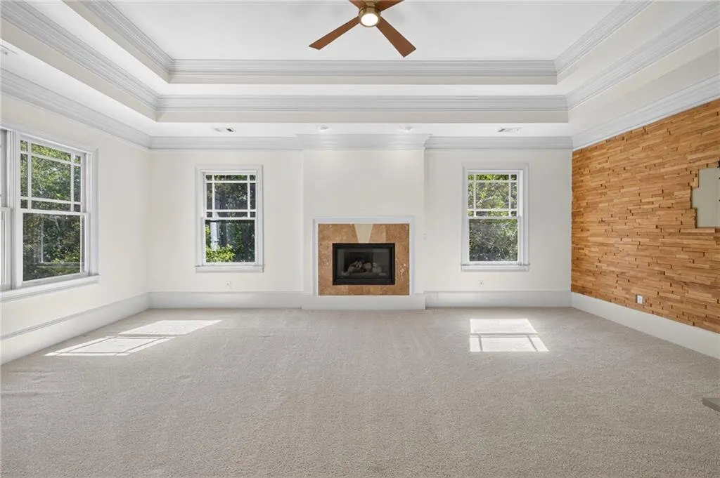 Tray ceiling with crown molding, fireplace with textured stone surround, wood-slat accent wall, and ceiling fan