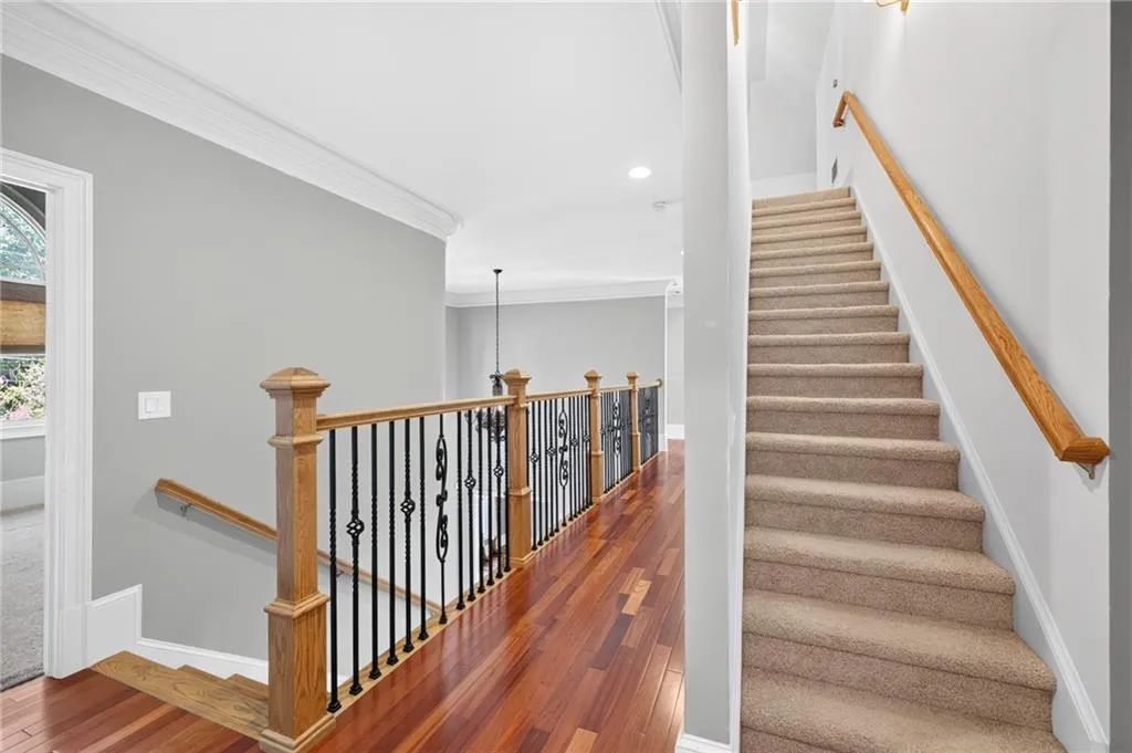 Staircase featuring carpeted steps, a wood-finish handrail, and a decorative metal balustrade with wood newel posts, complemented by wood-finish flooring and crown molding