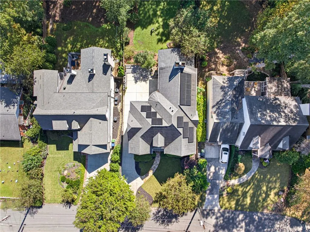 Aerial perspective of residential property featuring a shingled roof, roof-mounted solar panel arrays, a concrete driveway, and landscaped grounds with mature trees