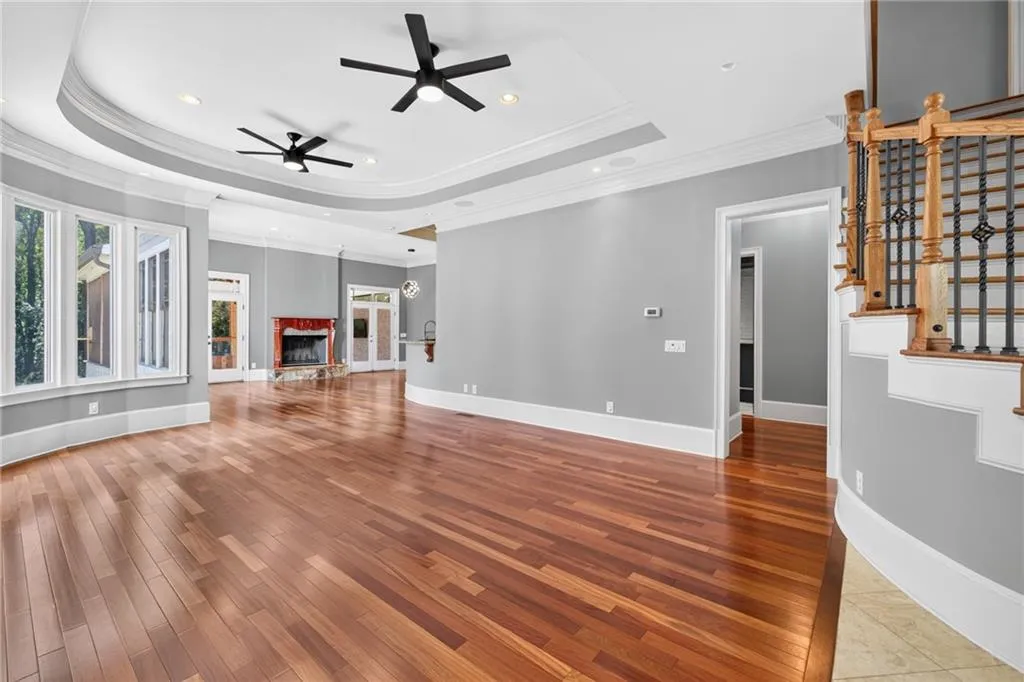Open-concept living space featuring wood-finish flooring, a tray ceiling, dual ceiling fans, a fireplace with a decorative surround, and a staircase with wood railings and metal balusters