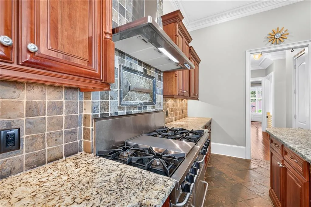 Kitchen featuring natural stone countertops, custom wood cabinetry, stainless steel gas range with griddle, stainless steel range hood, and stone tile backsplash
