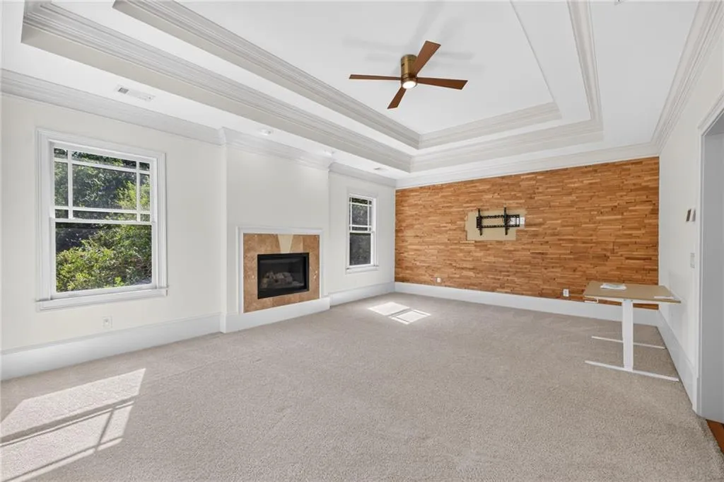 Tray ceiling with crown molding, natural wood-finished accent wall, tiled fireplace surround, and ceiling fan with integrated lighting