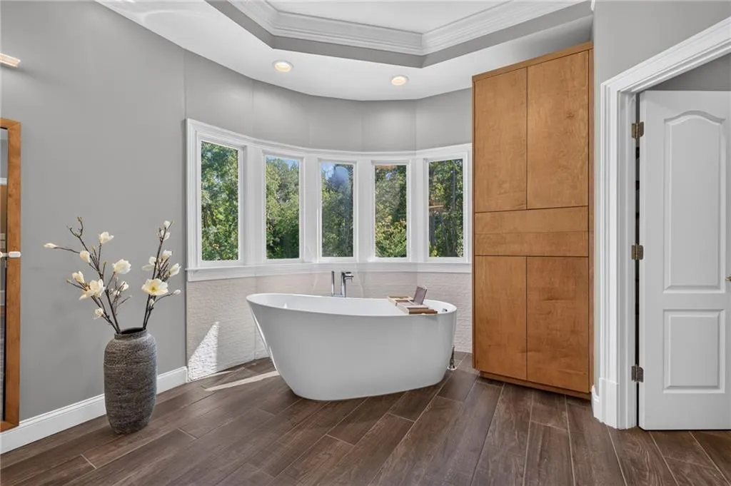 Freestanding soaking tub with a floor-mounted faucet, wood-finish flooring, built-in floor-to-ceiling cabinetry, recessed lighting, and a multi-panel bay window