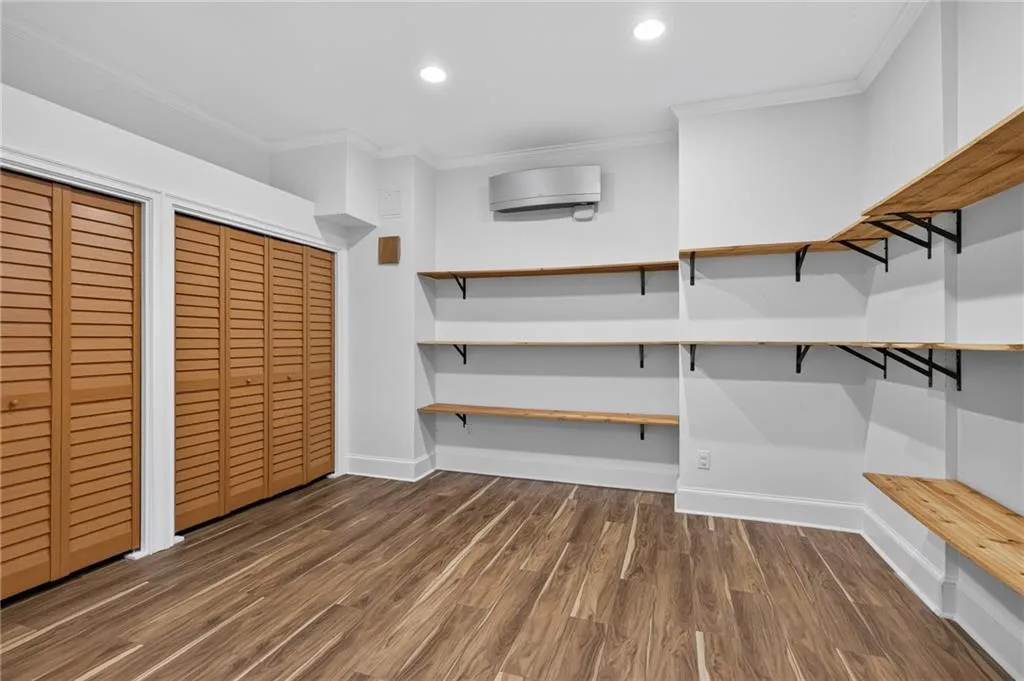 Wood-finish flooring, dual sets of louvered closet doors, wall-mounted ductless climate control unit, and custom natural wood shelving with black metal brackets