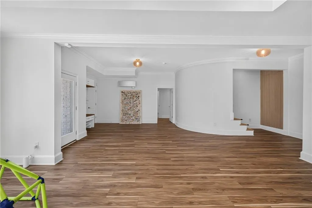 Open-concept interior featuring wood-finish flooring, curved architectural wall detail, built-in cabinetry, crown molding, and wall-mounted HVAC unit