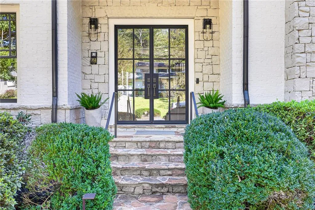 Light-toned stacked stone exterior featuring a double-entry door with a black metal grid frame, flagstone masonry steps, and exterior wall-mounted lanterns