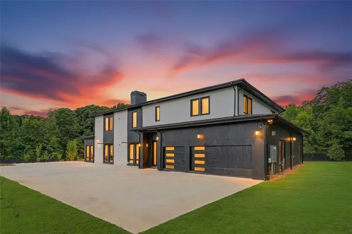 Contemporary exterior featuring a multi-tone facade, expansive concrete driveway, three modern garage doors with horizontal window inserts, mature trees bordering the property, and a manicured lawn