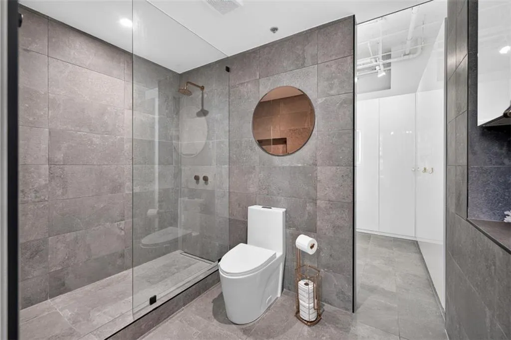 Full-height glass shower enclosure with bronze-finish shower fixtures, gray tile walls and flooring, and a round wall-mounted mirror
