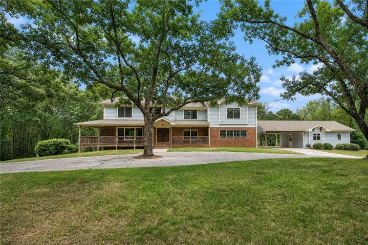 Expansive front yard featuring a paved drive, mature trees, a full-width front porch, light blue siding, and a brick facade