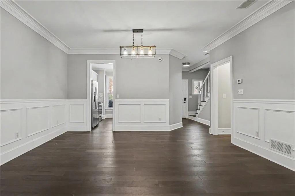 Unfurnished dining area featuring crown molding, dark wood-style flooring, a decorative wall, a wainscoted wall, and hanging lights