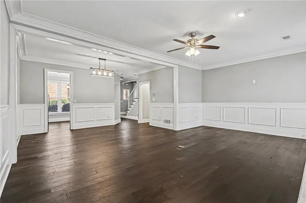 Unfurnished living room with a wainscoted wall, dark wood-style flooring, a ceiling fan, a decorative wall, and crown molding