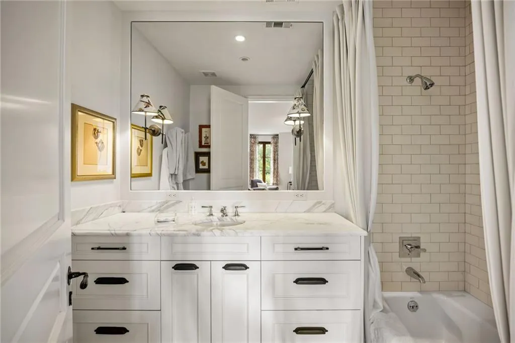 White vanity with dark hardware, stone countertop, integrated dual sinks, large wall-mounted mirror, and subway tile shower surround