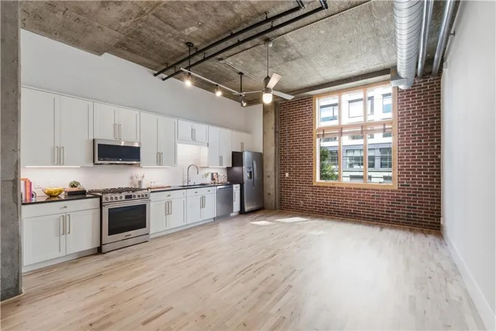 Exposed concrete ceiling with industrial track lighting, light wood-finish flooring, white flat-panel cabinetry, stainless steel appliances, and a brick accent wall with a large window