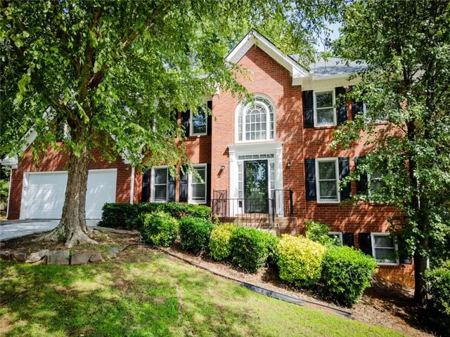 Traditional brick facade residence featuring a prominent arched window, a two-car garage, and a manicured sloped lawn with mature trees