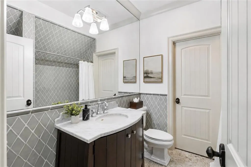 Full-width vanity mirror, dark wood-finish vanity with white countertop, integrated sink with chrome fixtures, tiled shower with a curtain rod, and a white toilet