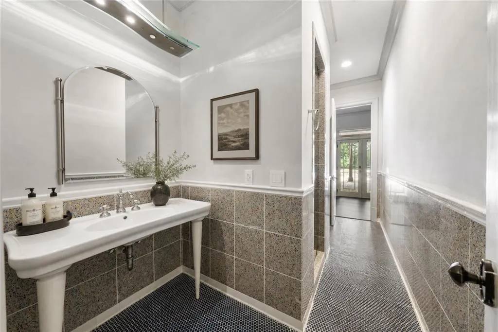 Pedestal sink with polished chrome fixtures, arched vanity mirror, mosaic floor tiling, and wainscoting
