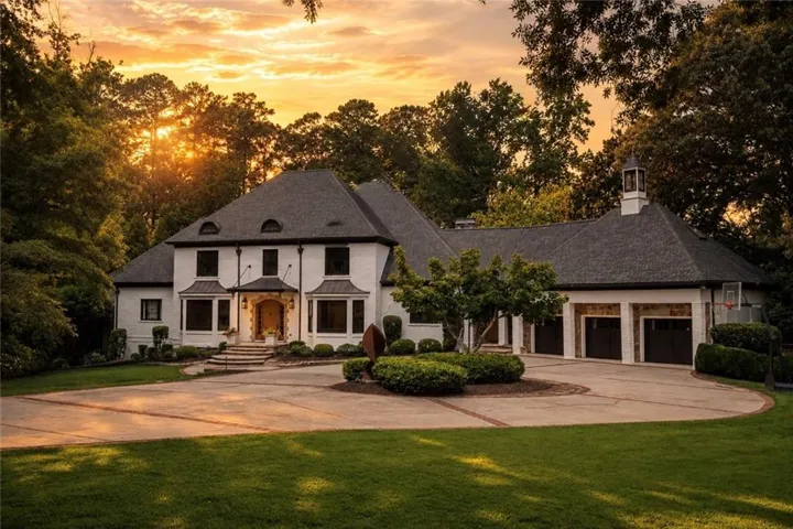 Circular driveway with brick accents, white stucco exterior, multiple garage bays with wood-finish doors, cupola feature, and mature landscaping