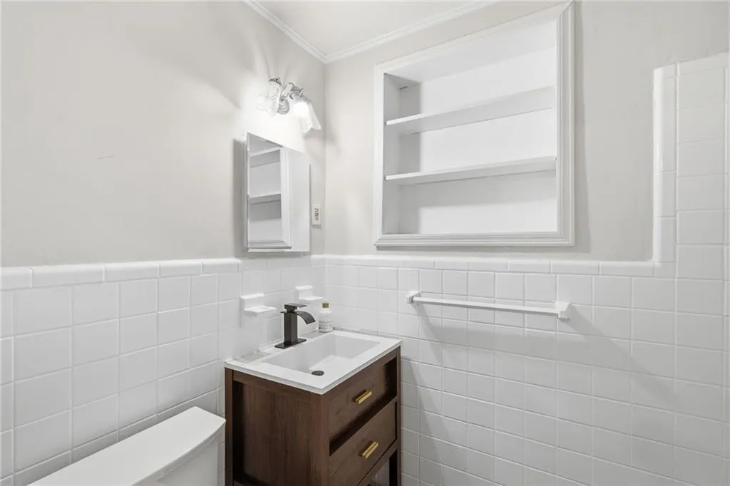 White tile wainscoting, recessed shelving, wall-mounted light fixture, wood-finish vanity with drawer storage, and a basin sink with a black faucet