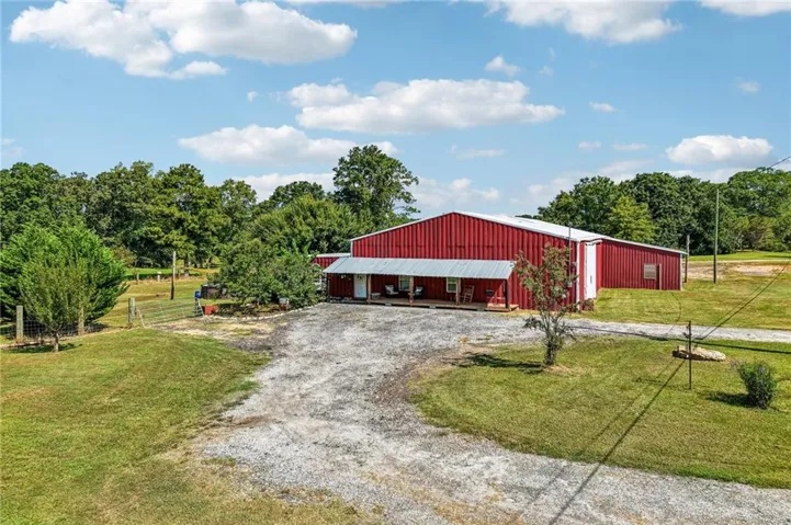 Red metal-sided structure with a white metal roof, featuring a covered front porch, gravel drive, and surrounding mature trees