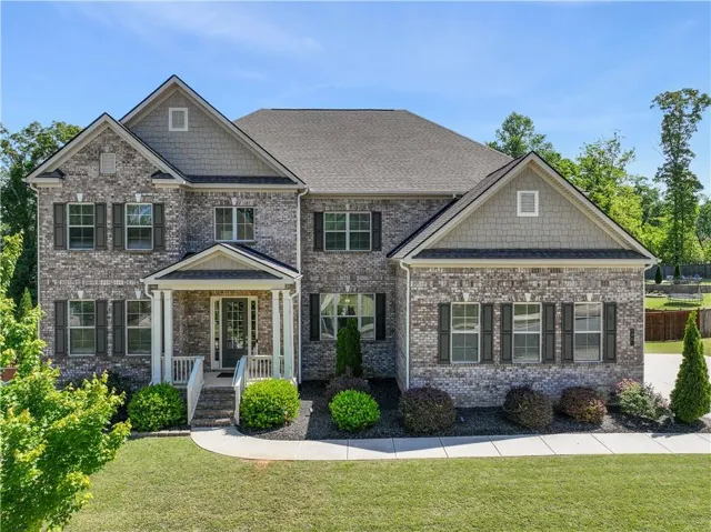 Traditional two-story residence featuring a brick facade with shingle accents, dark shutters, a covered front porch with white columns, and a landscaped front yard with a concrete pathway
