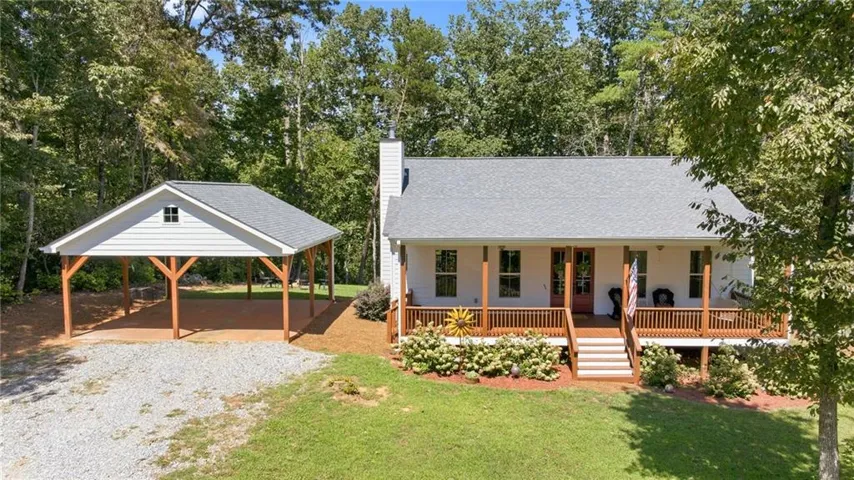 White exterior with a covered front porch, wood porch railing, exterior steps, and a detached carport