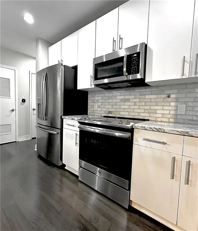 Kitchen featuring appliances with stainless steel finishes, backsplash, light stone countertops, dark wood-style flooring, and white cabinets