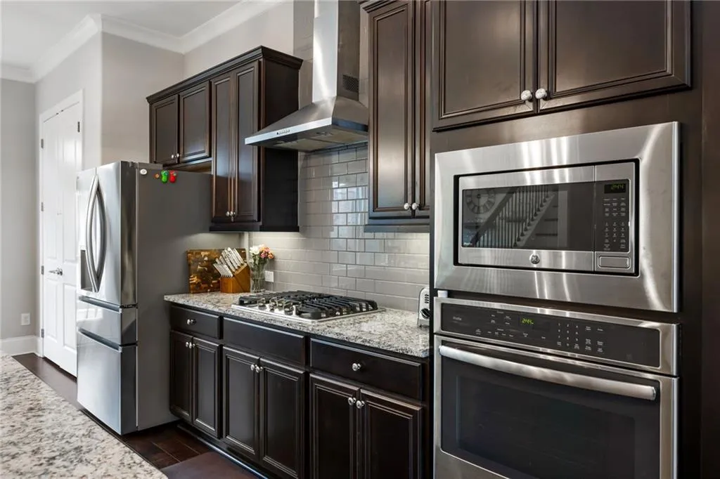 Kitchen featuring tasteful backsplash, appliances with stainless steel finishes, wall chimney range hood, light stone countertops, and dark wood-style floors