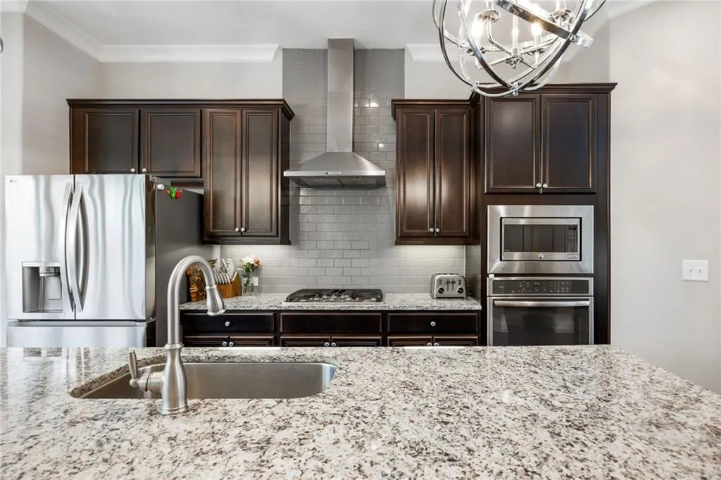 Kitchen featuring dark brown cabinets, tasteful backsplash, stainless steel appliances, wall chimney range hood, and light stone counters