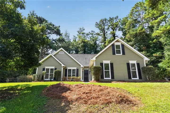 Front exterior featuring green siding, black shutters, dormer window, and mature trees