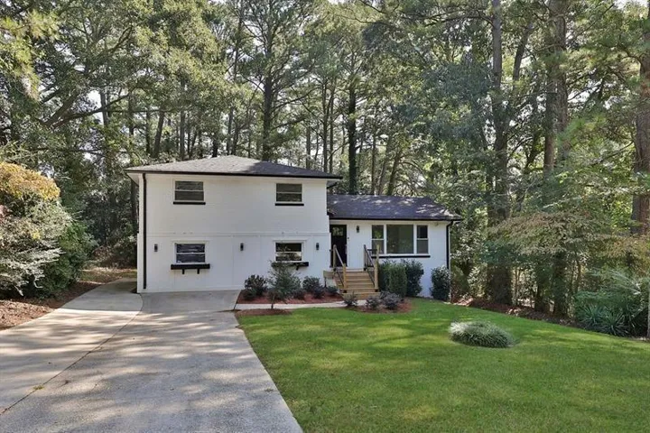 White exterior with black trim, concrete driveway, front lawn, mature trees, and natural wood stairs