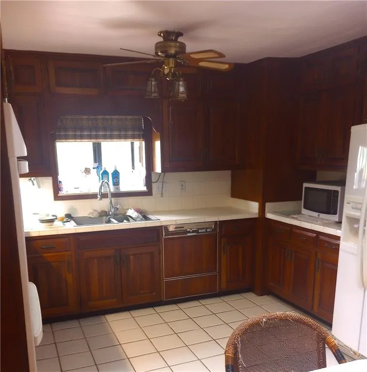 Kitchen with tile countertops, light tile patterned floors, a ceiling fan, paneled dishwasher, and white refrigerator with ice dispenser
