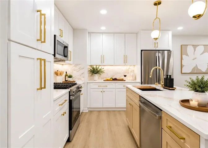 Modern kitchen featuring an island with integrated sink, stainless steel appliances, white cabinetry with gold-tone hardware, a marbled backsplash, and wood-finish flooring