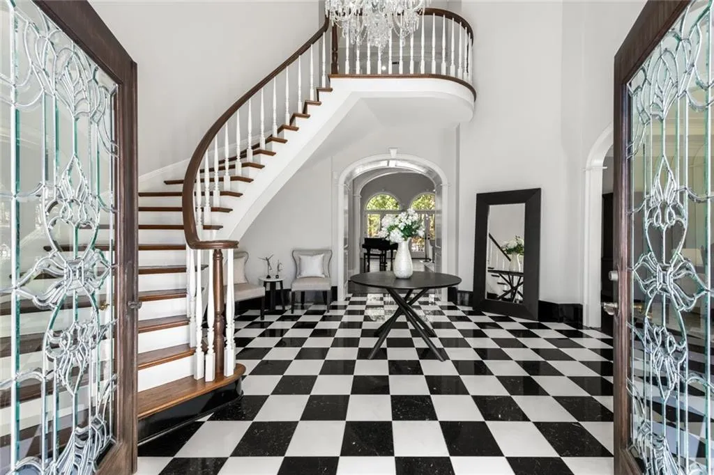 A dramatic two-story foyer makes a stunning first impression, featuring a sweeping curved staircase and striking black-and-white checkered marble flooring. Elegant iron double doors and abundant natural light enhance the grand, open feel of this sophisticated entry space.
