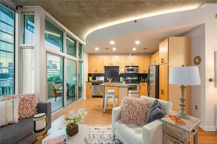 Open concept living space featuring wood-finish flooring, a full kitchen with light wood cabinetry and stainless steel appliances, a kitchen island with seating, and a balcony with railings