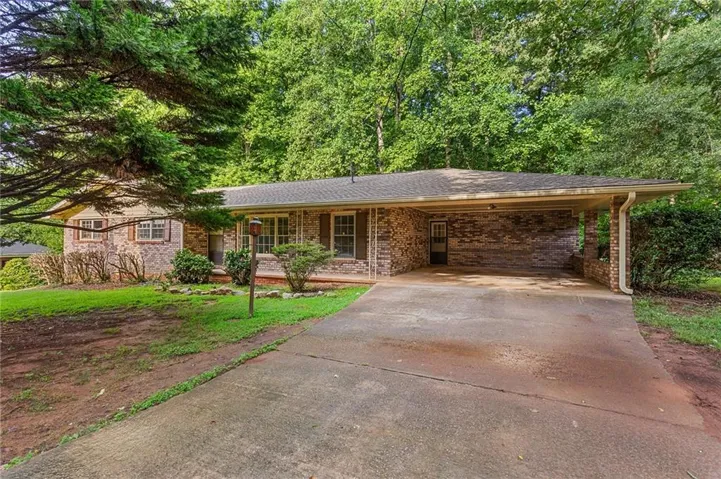 Brick facade, covered carport, concrete driveway, lush green landscaping, and mature trees
