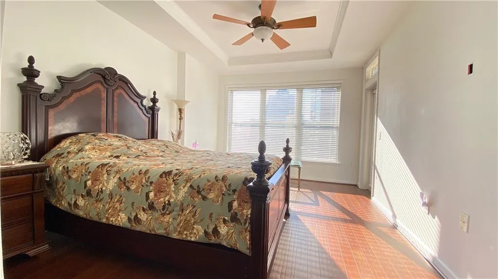 Recessed tray ceiling with a built-in ceiling fan, expansive window with blinds, and wood-finish flooring