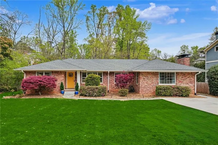 Brick exterior with a gray shingle roof, featuring a bright yellow front door, a driveway, and mature landscaping