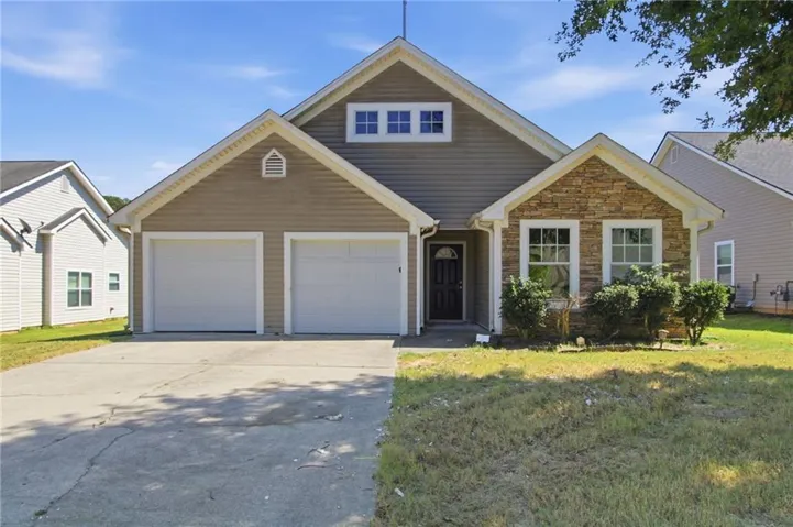 Welcoming two-car garage with white panel doors, stone and siding exterior combining warm earth tones, arched roofline with gabled dormers and upper-level window detail, manicured front lawn with mature landscaping, and a concrete driveway