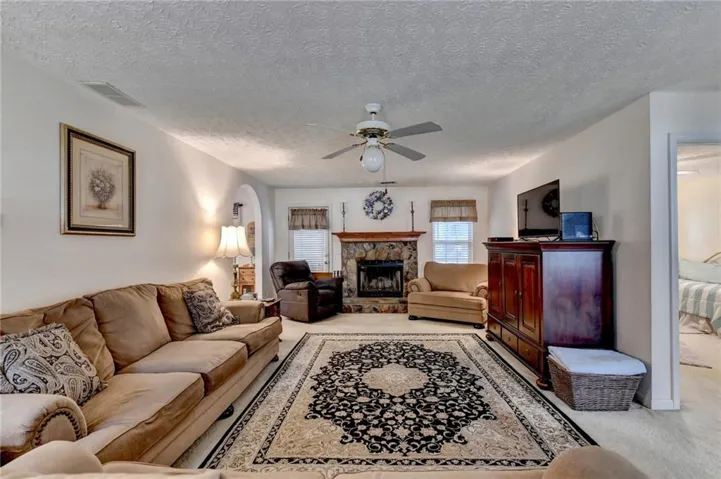 Living area featuring a stone-faced fireplace with a wood mantel, a ceiling fan with integrated lighting, and light-colored carpeting