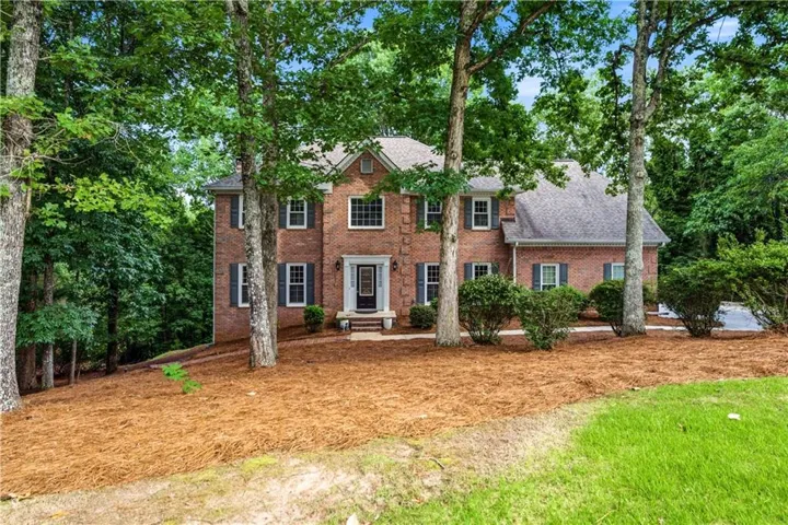 Brick facade residence featuring a front entry portico, multiple windows with shutters, a gabled roofline, and mature trees surrounding the property