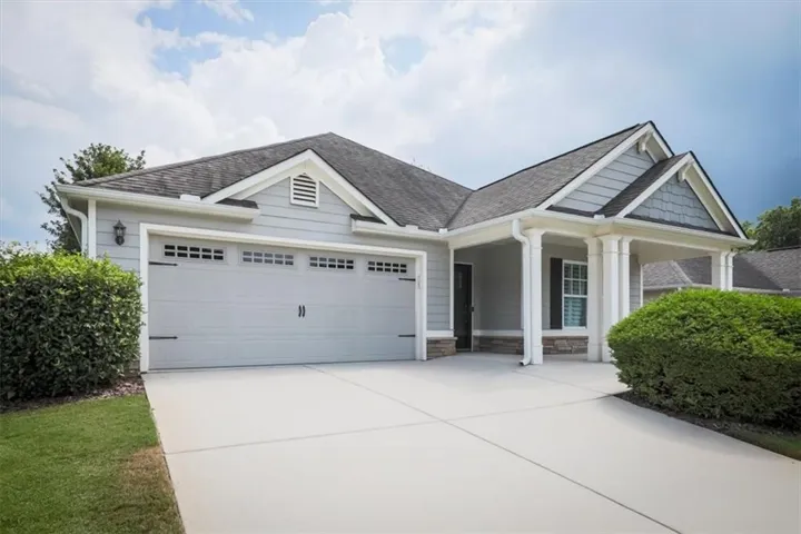Exterior featuring light gray siding, a dark asphalt shingle roof, a two-car garage with decorative hardware, a covered front entry with white columns, and stone accents