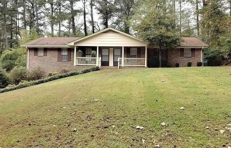 Brick-clad residence featuring a covered front porch with white railing, brown shutters, and a sloped front lawn