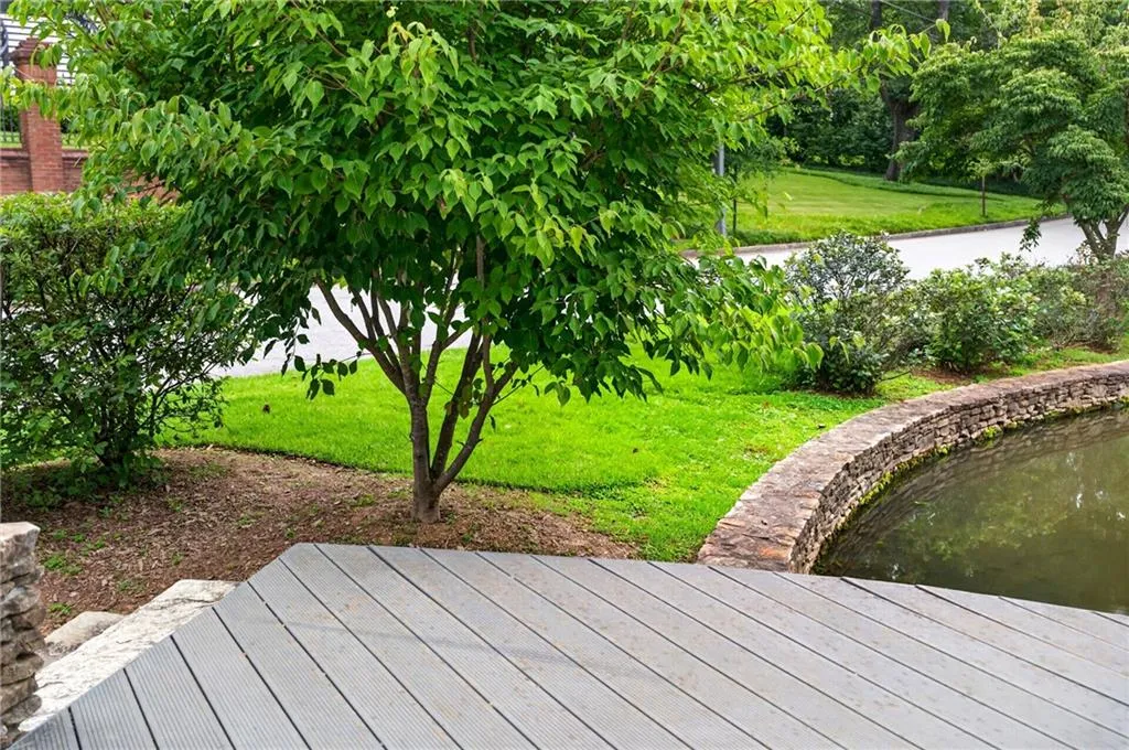 Composite decking with a stone retaining wall bordering a water feature, surrounded by mature landscaping and a lush lawn