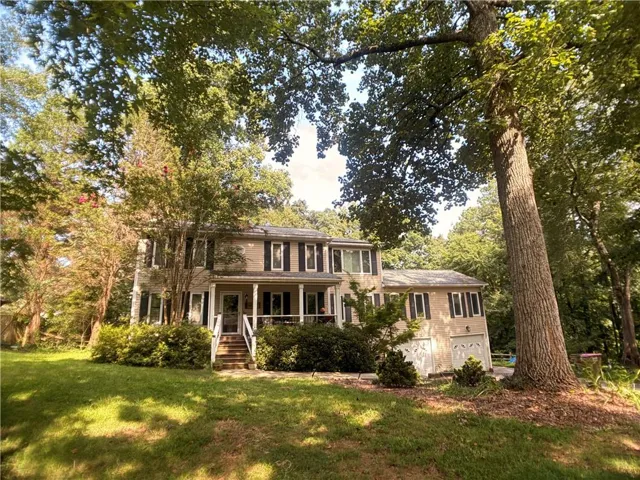 Two-story residence featuring a covered front porch, dark shutters, an attached garage, and a sloped front lawn with mature trees