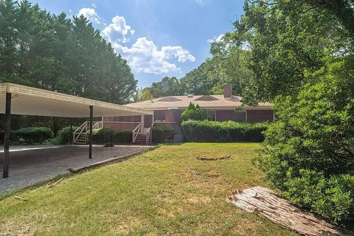 Carport with covered parking, brick exterior, multiple rooflines, a prominent chimney, and mature perimeter trees