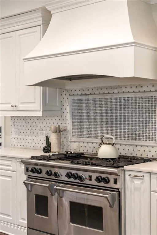 Kitchen featuring premium range hood, range with two ovens, white cabinets, backsplash, and light stone counters