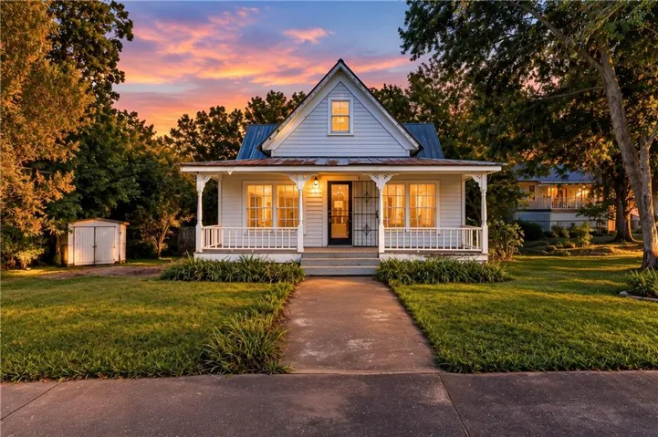White clapboard siding, covered front porch with white railing, centered front door with security screen, mature trees, and a detached shed